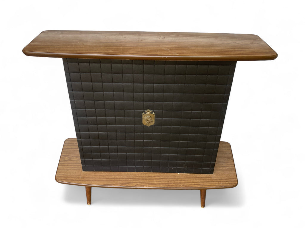 A mid century bar with storage shelves in the rear. Laminated top and foot rest. Faux leather with a crest in the middle.